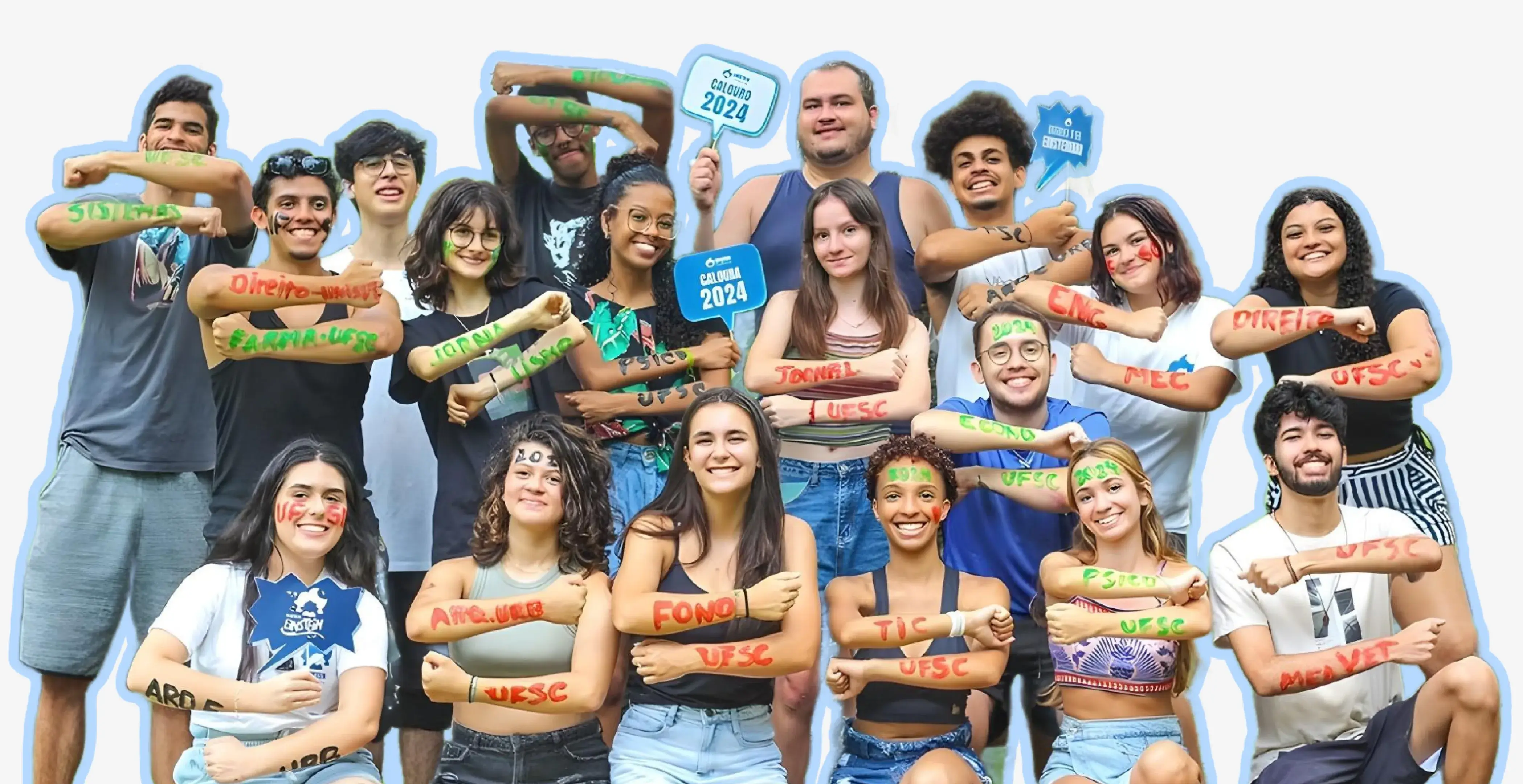 Grupo de estudantes calouros de 2024, posando para foto em clima de celebração. Todos têm os nomes dos cursos escritos nos braços, como 'Sistemas', 'Psico', 'Jornal', entre outros, e alguns seguram placas com as palavras 'Calouro 2024' e 'UFSC'. A expressão deles é de alegria e animação.